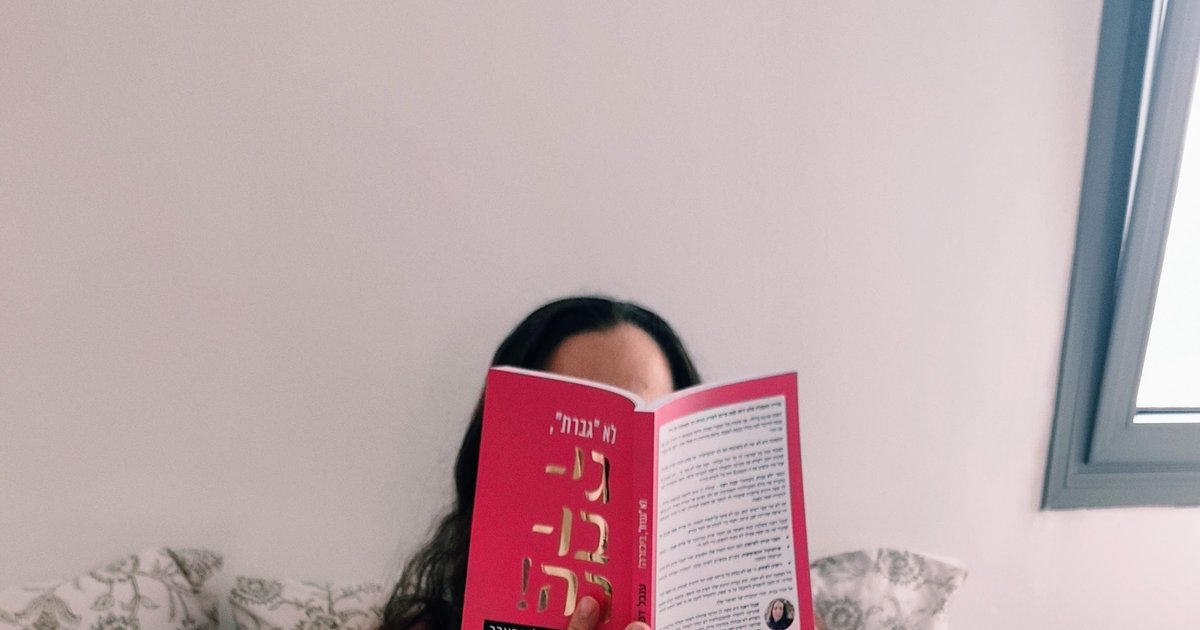 אישה שוכבת במיטה וקוראת ספר בכריכה ורודה, לצידה כלב קטן בצבע לבן-בז'
